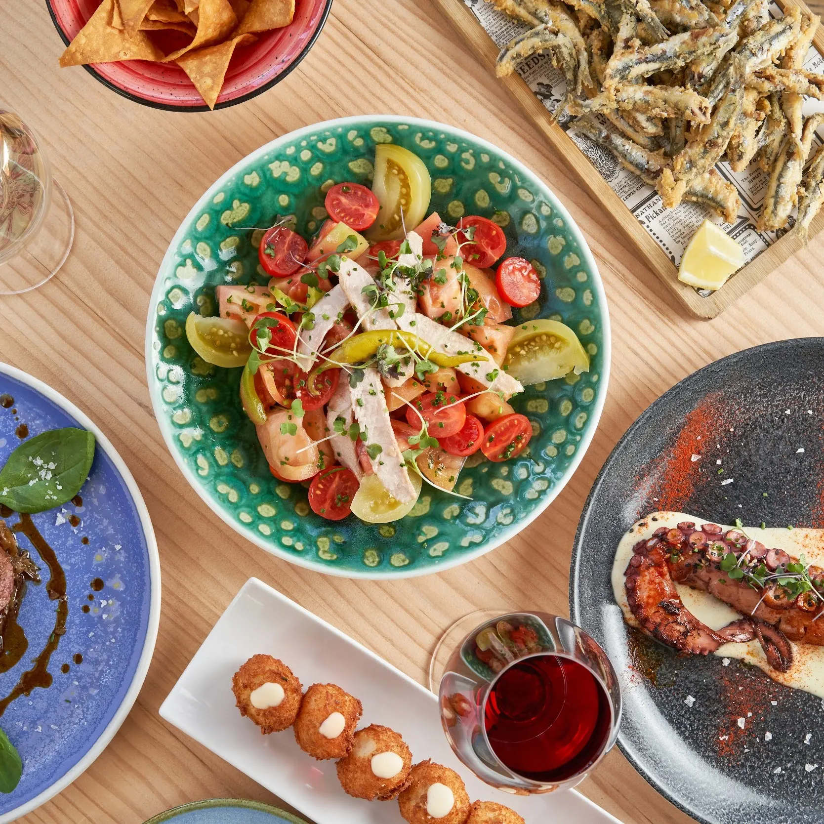 La imagen muestra una variedad de platos del Maná 75. Hay un plato con un filete adornado con hierbas y salsa, otro plato con pulpo a la parrilla acompañado de rodajas de limón y una salsa cremosa, y un bol con una ensalada de tomates, cebollas.