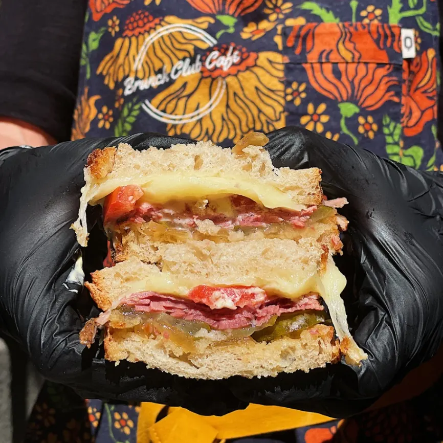 Persona con guantes negros sostiene sándwich con queso fundido, carne y verduras; lleva delantal floral de “Denver Club Café”.