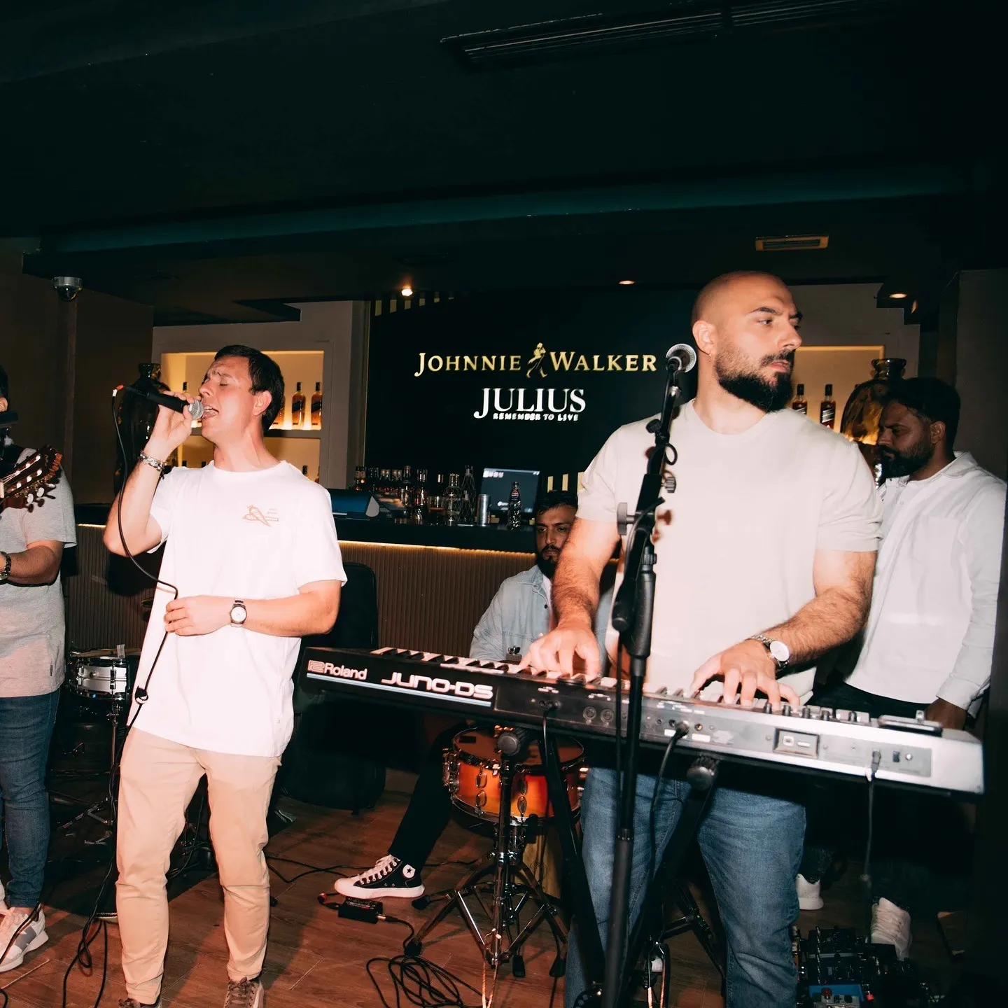 Concierto en vivo en Julius con banda tocando y cantante al micrófono. Ambiente animado y barra con visibilidad de Johnnie Walker.