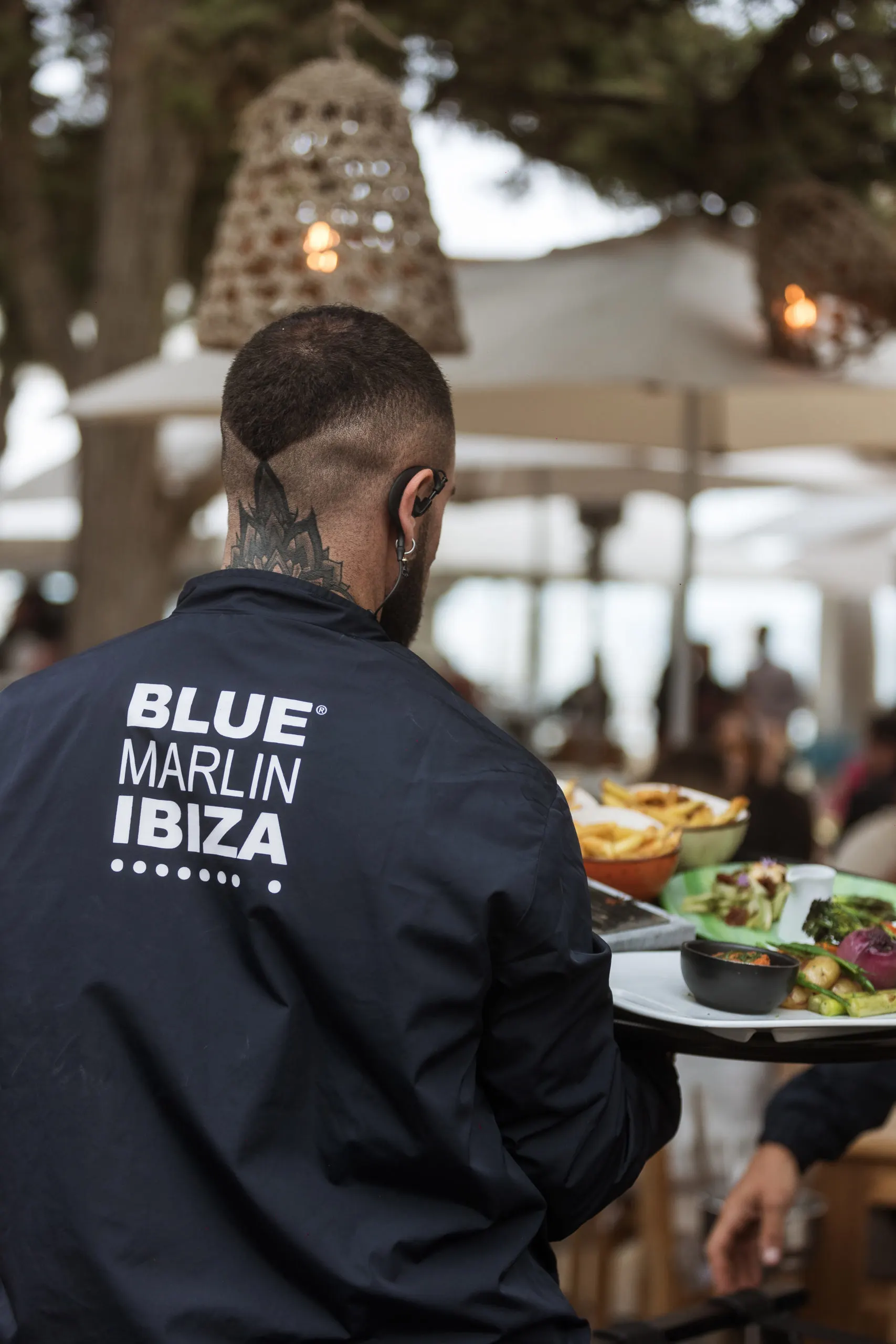 Camarero con tatuaje sirve platos en Blue Marlin Ibiza, reflejando estilo, atención personalizada y ambiente exclusivo junto al mar.
