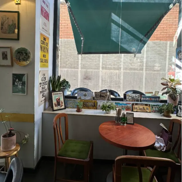 Cafetería acogedora con mesa redonda de madera, sillas verdes, ventana con plantas y carteles decorativos de cócteles en pared iluminada.