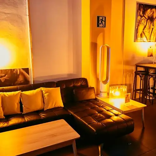Sala con sofá negro, luz cálida, mesa de madera y póster en pared. Ambiente acogedor con decoración íntima y mesas altas al fondo.