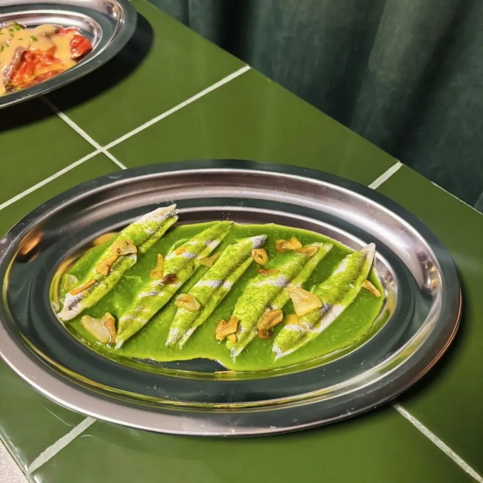 Filetes de pescado en salsa verde con ajo sobre plato ovalado de acero; fondo con azulejos verdes y otros platos parcialmente visibles.
