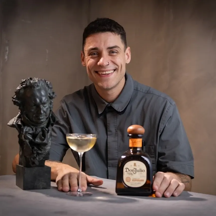 Manuel Spolaore with a cocktail and bottle in hand, smiling at the camera, while sitting at a table.