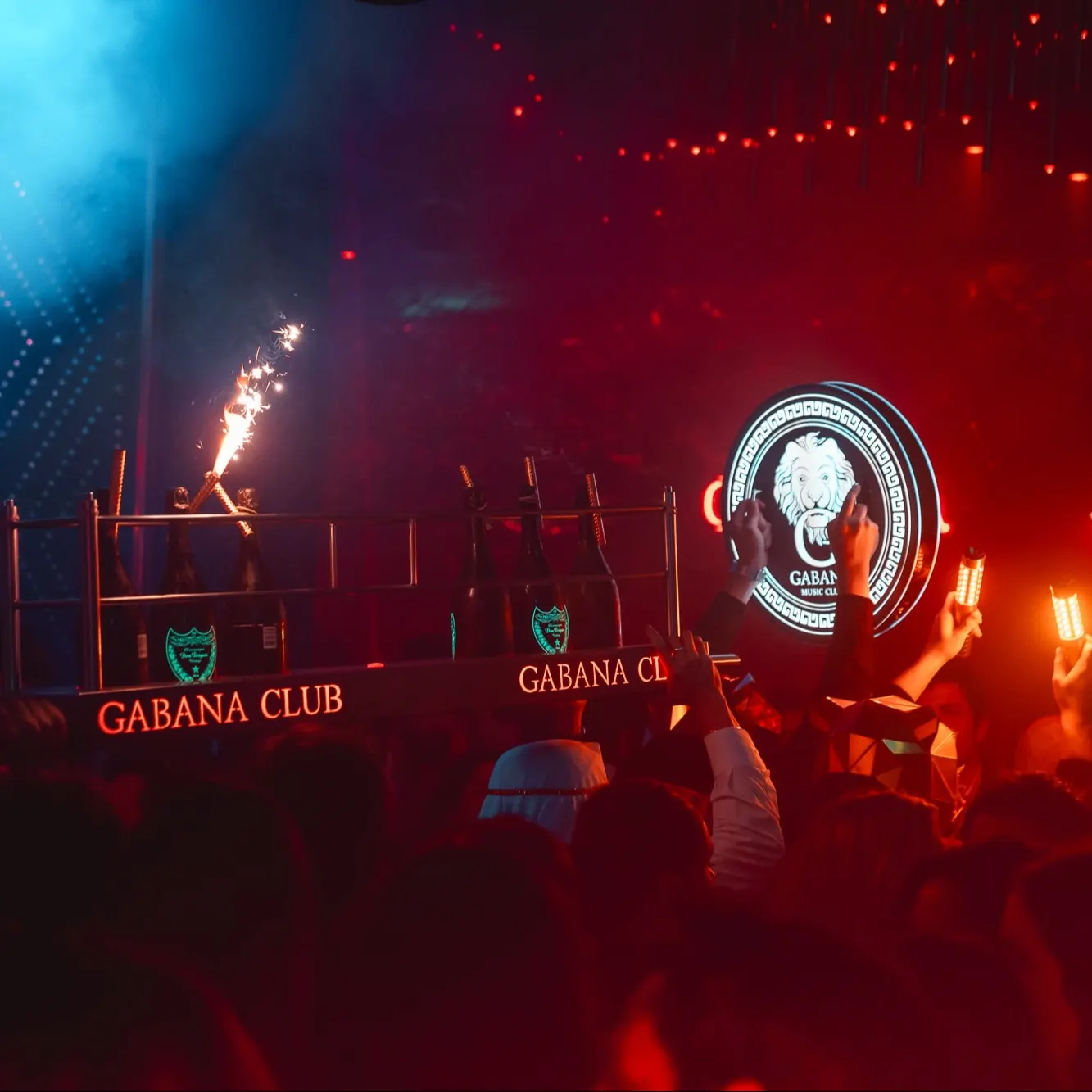 Gabana: Ambiente de fiesta en Gabana Club con luces rojas y botellas en bandeja iluminada con bengalas, rodeadas de una multitud animada. El logo del club destaca en el fondo.