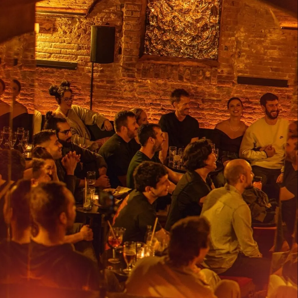 Grupo de personas en bar con paredes de ladrillo, luz tenue y ambiente íntimo; bebidas en mano y altavoz en pared para música o presentaciones.