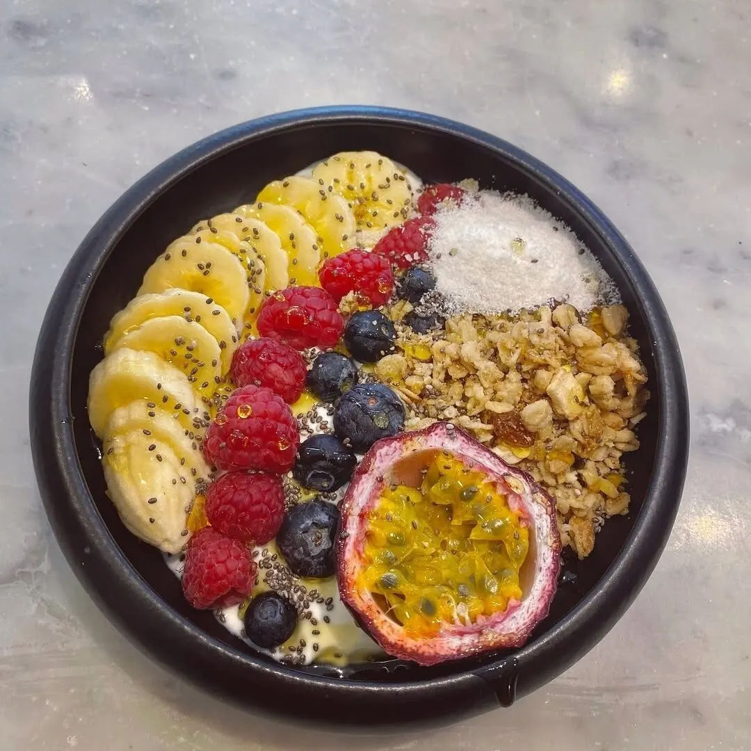 Bol de yogur con plátano, frambuesas, arándanos, granola, chía, coco rallado y medio maracuyá sobre superficie de mármol.