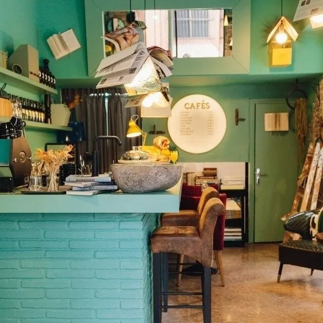 Cafetería acogedora con barra turquesa, lámparas de libros abiertos, menú de cafés, estanterías y zona de mesas cómodas.