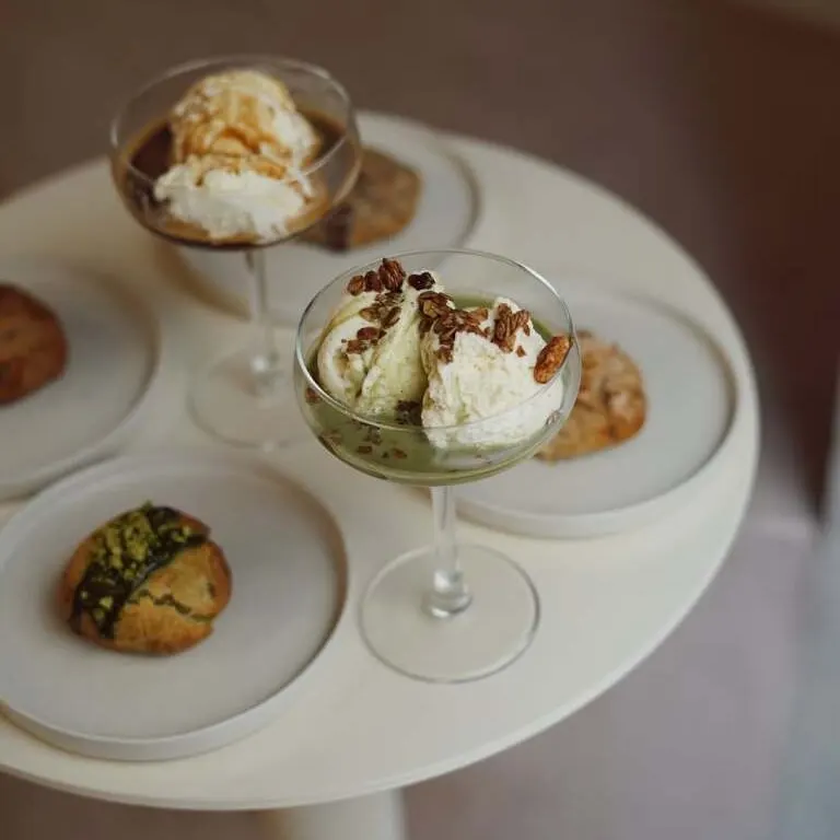 Postres servidos en copas con helado, sirope y frutos secos, acompañados de galletas y bollería sobre platos individuales en una mesa redonda blanca.
