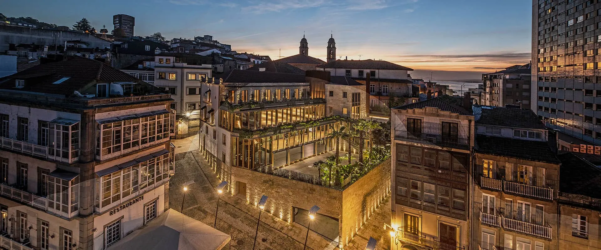 Imagen aérea del Palacio de la Oliva iluminado al anochecer, rodeado de edificios históricos y con el mar al fondo.