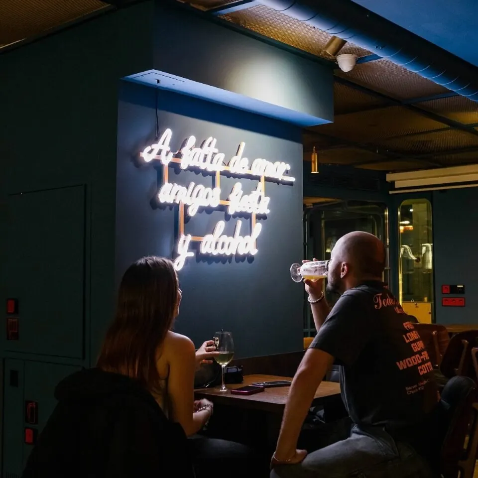 Dos personas sentadas en mesa de bar con luz tenue. Al fondo, neón encendido con la frase: “A falta de amor, amigos, fiesta y alcohol.”