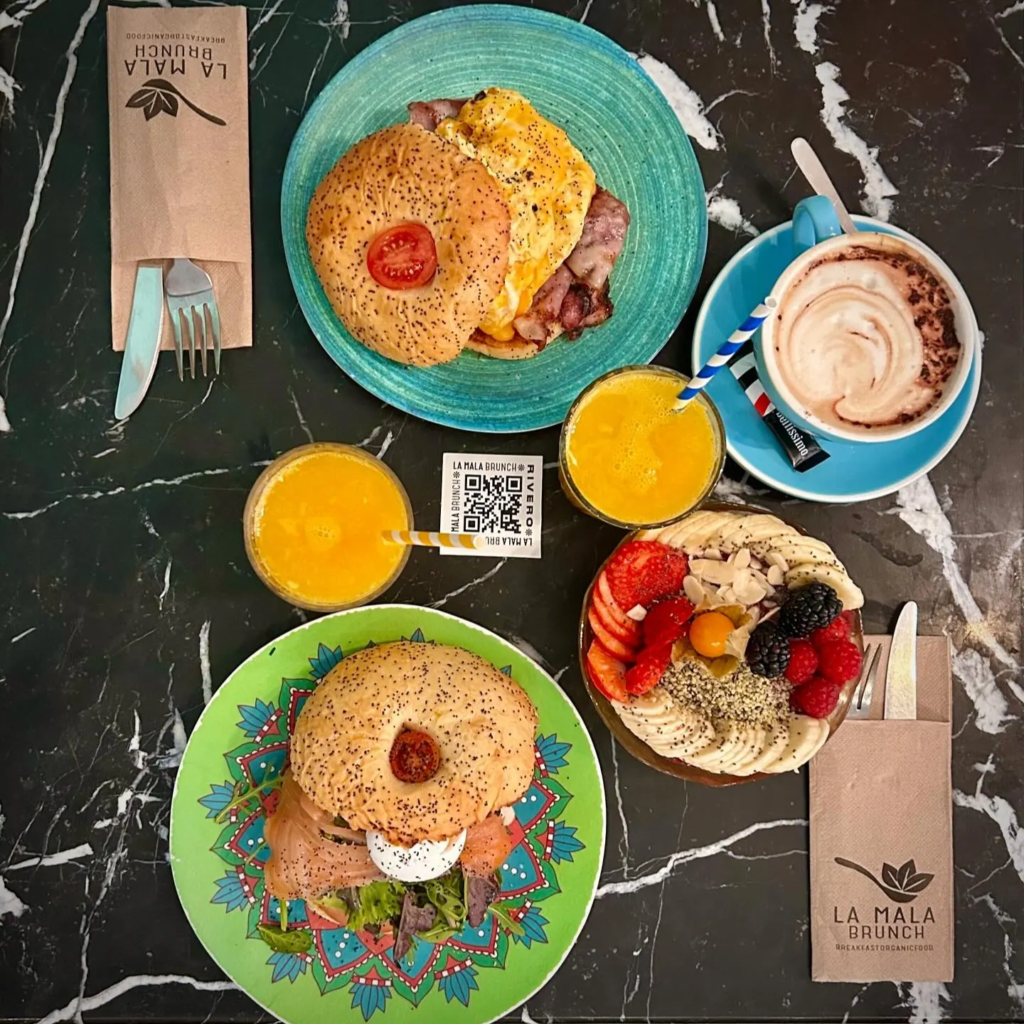 Desayuno en La Mala Brunch con bagels, jugo de naranja, tazón de frutas y chocolate caliente servido sobre mesa de mármol.