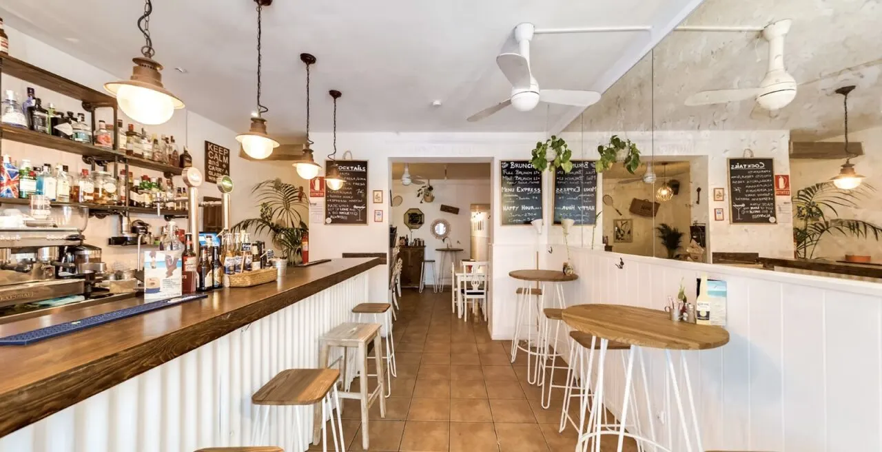 Interior de bar con barra de madera, taburetes altos, pizarras con menús escritos a mano y decoración luminosa con ventiladores y plantas colgantes.