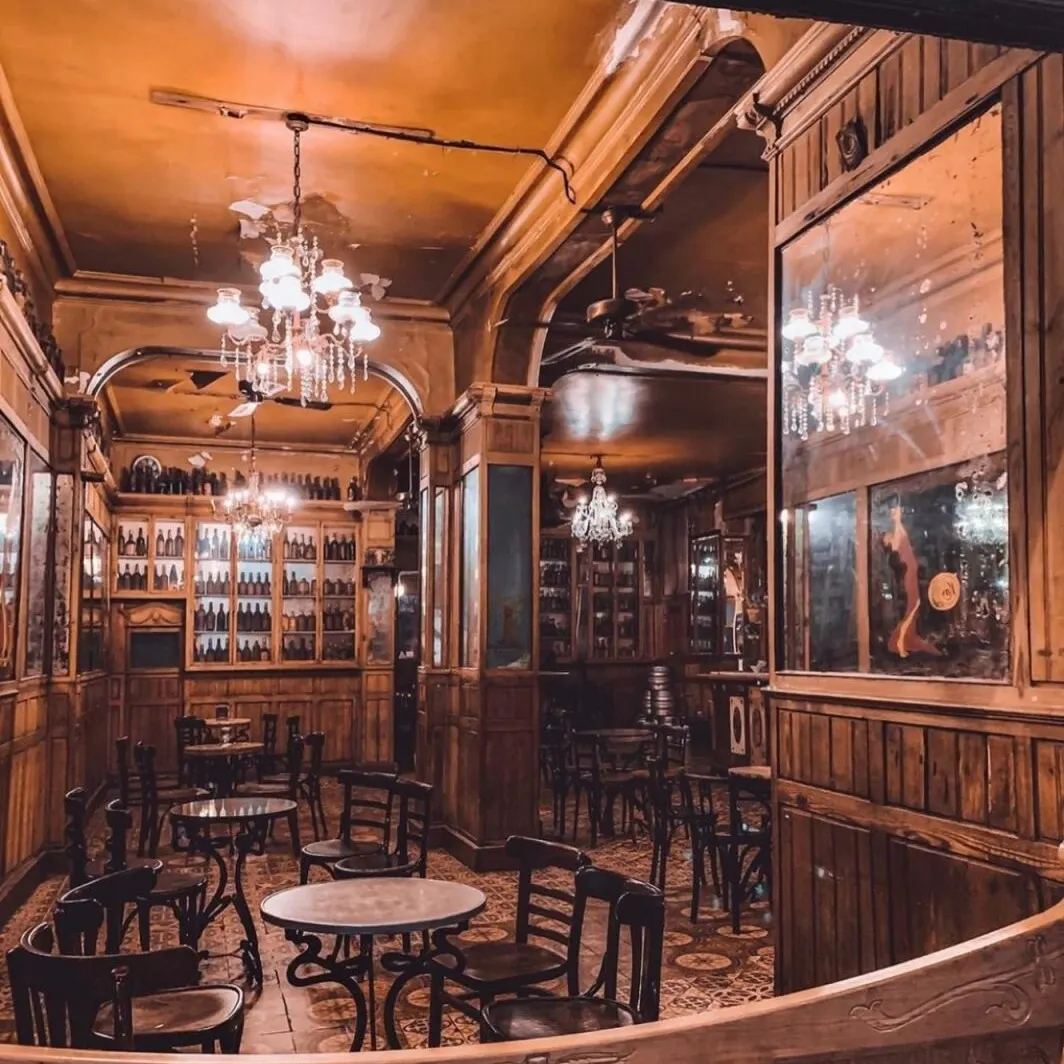 Café vintage con mesas redondas, estanterías con botellas, lámparas de araña y ambiente cálido y nostálgico.