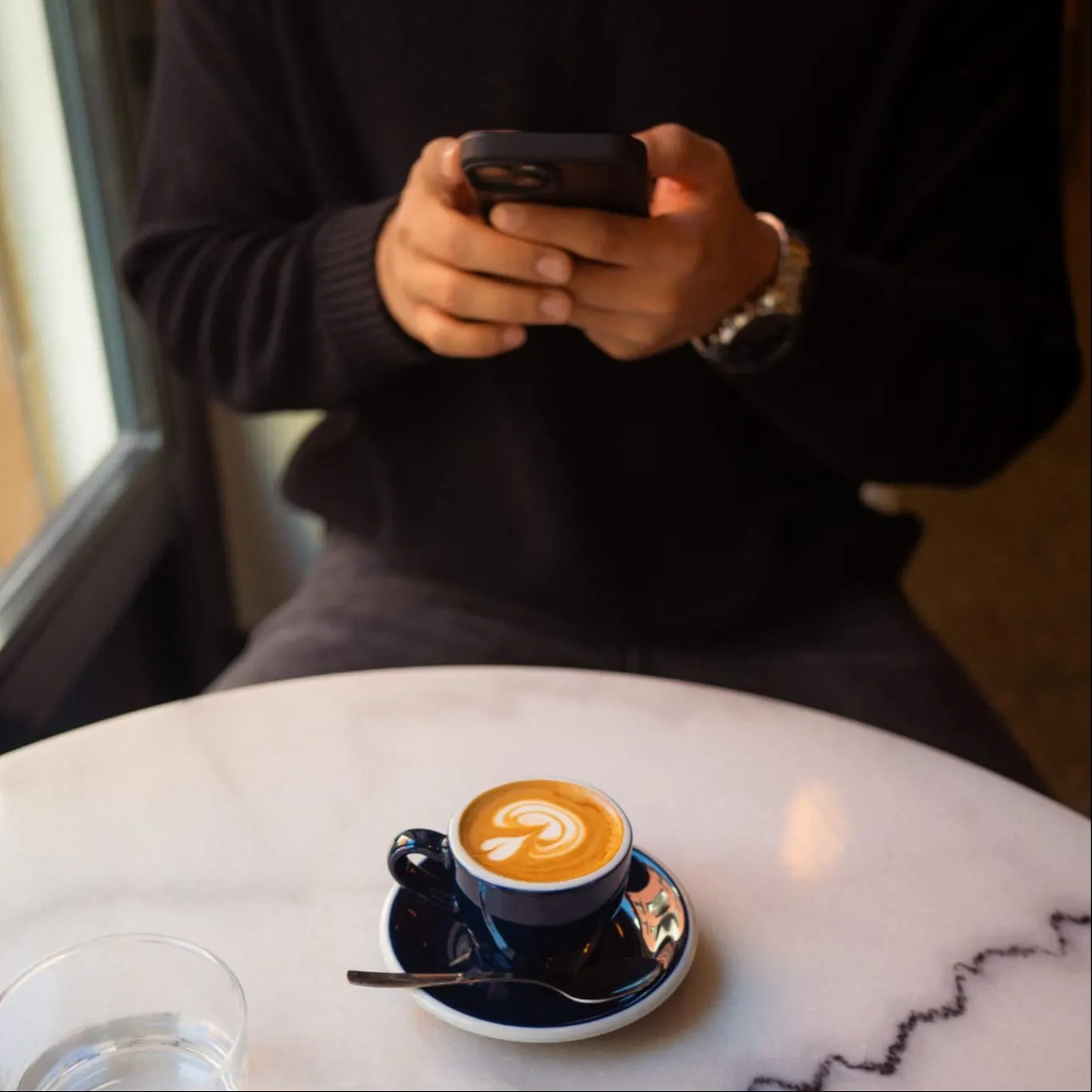 Café con arte latte servido en taza azul sobre mesa de mármol mientras una persona revisa su móvil en Trava de Noche.