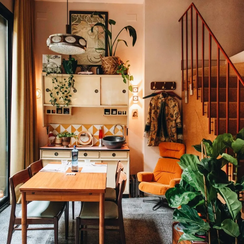 Comedor acogedor con mesa de madera, lámpara vintage, plantas, sillón naranja y escalera junto a ventana con cortinas beige.