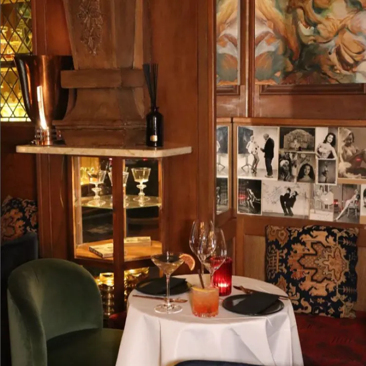 La imagen muestra un entorno de comedor íntimo de Nuts Indulgence. Hay una pequeña mesa redonda cubierta con un mantel blanco, con dos copas de vino, una de vino tinto y otra de vino blanco, y un cóctel. Las sillas de la mesa están en terciopelo