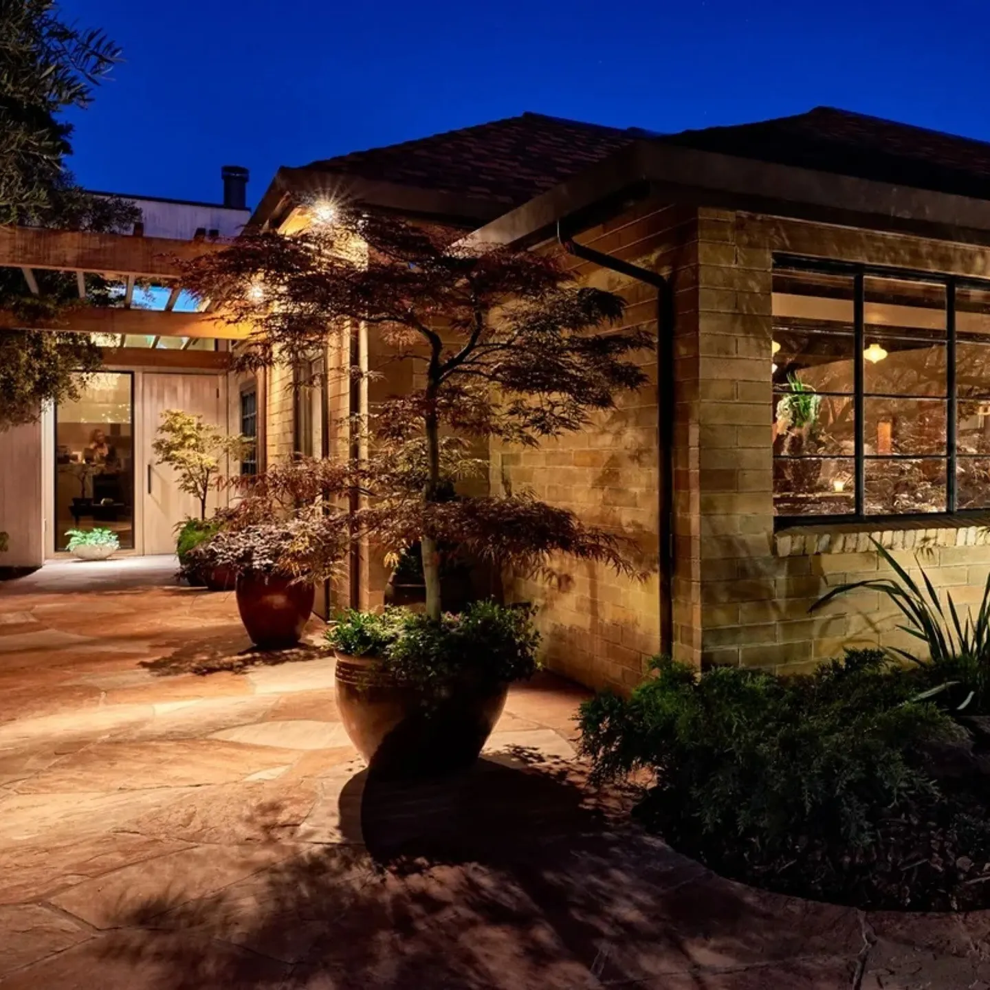 Entrada nocturna de Cenador de Amós, con iluminación cálida, vegetación cuidada y arquitectura rústica que invita a una experiencia elegante.