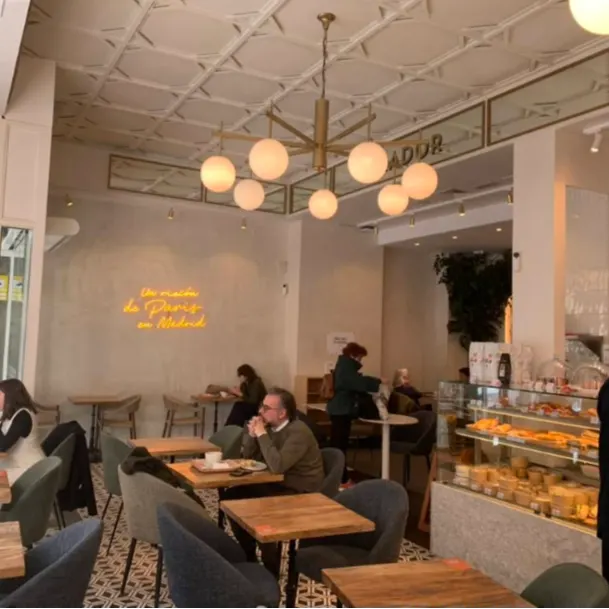 Interior de Maison Kayser Gaztambide con su característico neón "Un rincón de París en Madrid", decoración elegante y clientes disfrutando del café.