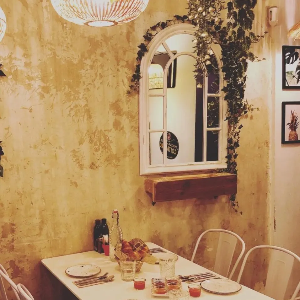 Comedor rústico con mesa para cuatro, pan, agua y salsa roja. Espejo arqueado, plantas colgantes y luces cálidas decoran el ambiente.