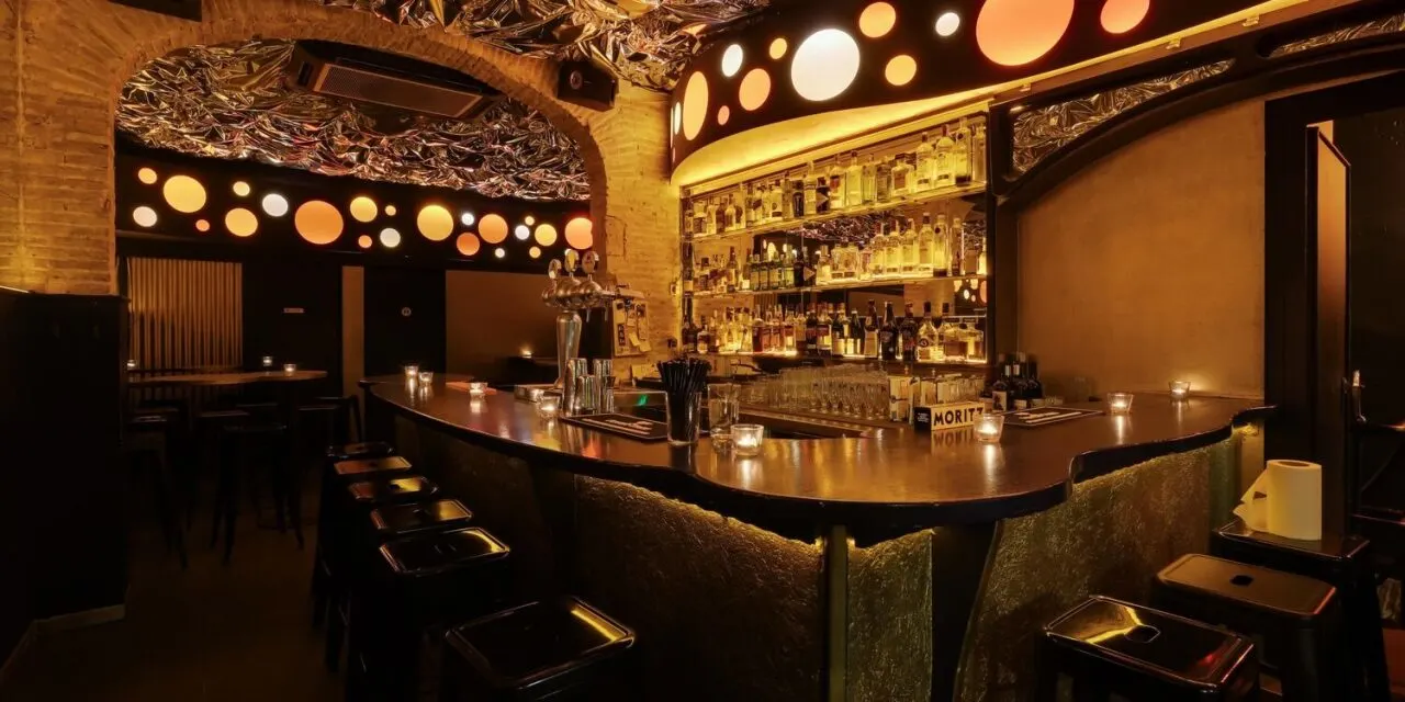 Bar moderno con barra curva, estanterías con botellas y luces circulares decorativas. Mesas al fondo y ambiente cálido y estilizado.