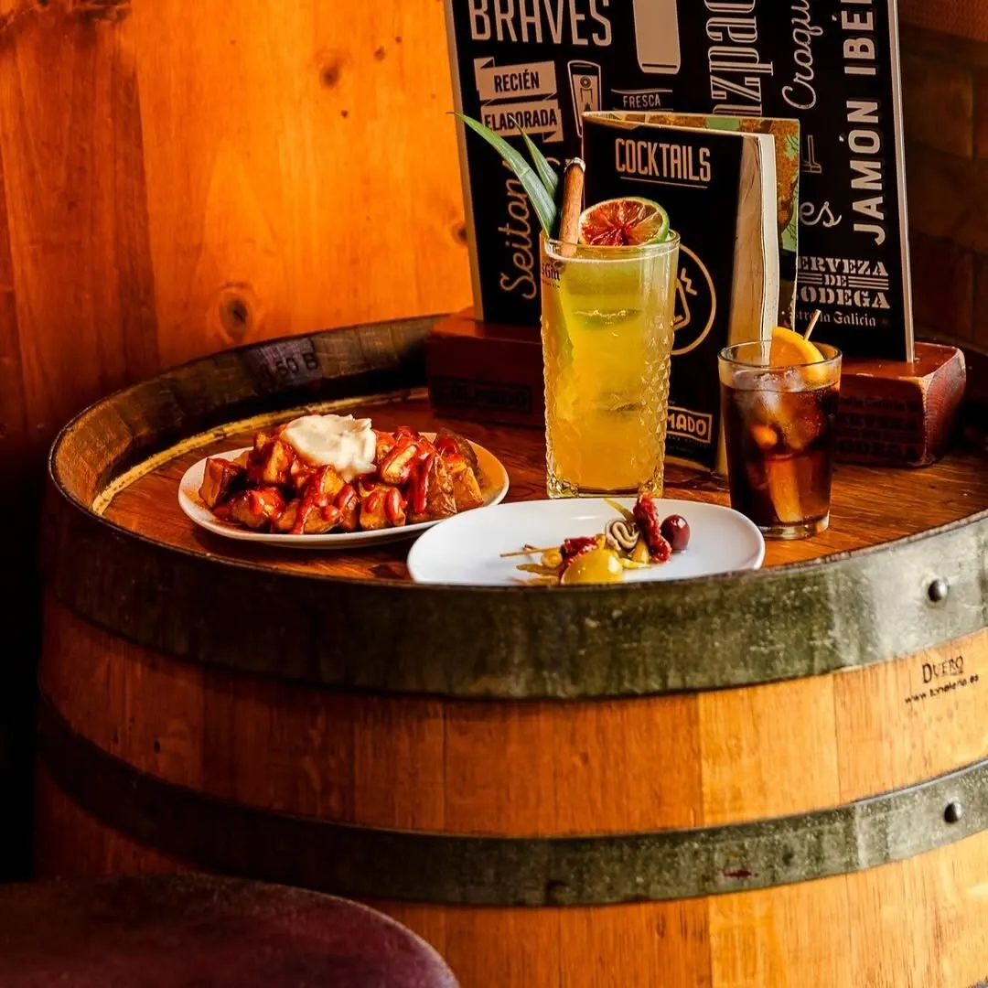 Barril de madera usado como mesa con cócteles amarillos y naranjas, tapas con pan, tomate y brochetas; fondo con cartel de palabras gastronómicas.