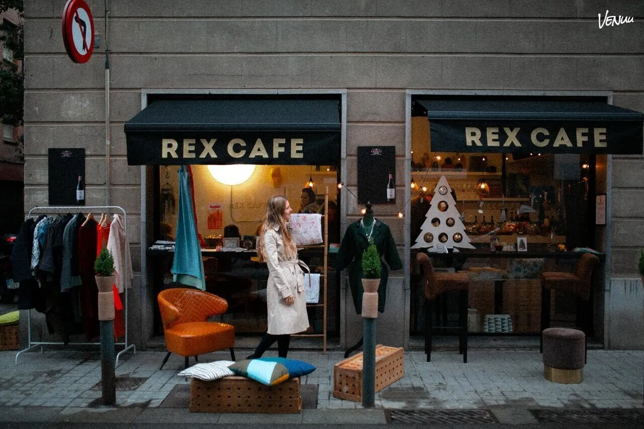 Fachada de Rex Café con toldos negros, decoración navideña, prendas expuestas en la calle y una mujer caminando frente al local.