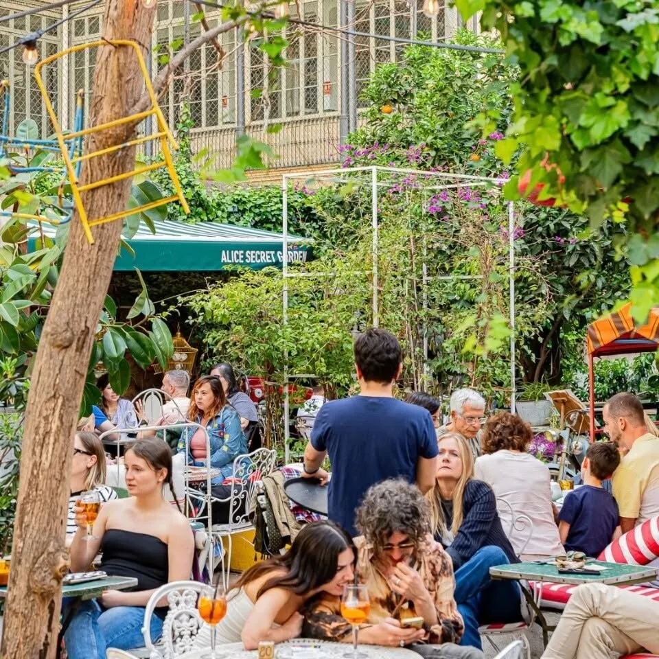 Café exterior lleno de gente bajo toldo verde “ALICE SECRET GARDEN”, rodeado de plantas y árboles que crean un ambiente frondoso y animado.