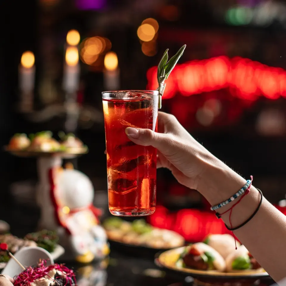 Mano levantando un cóctel rojo decorado con hoja verde, con platos de comida y luces desenfocadas al fondo.