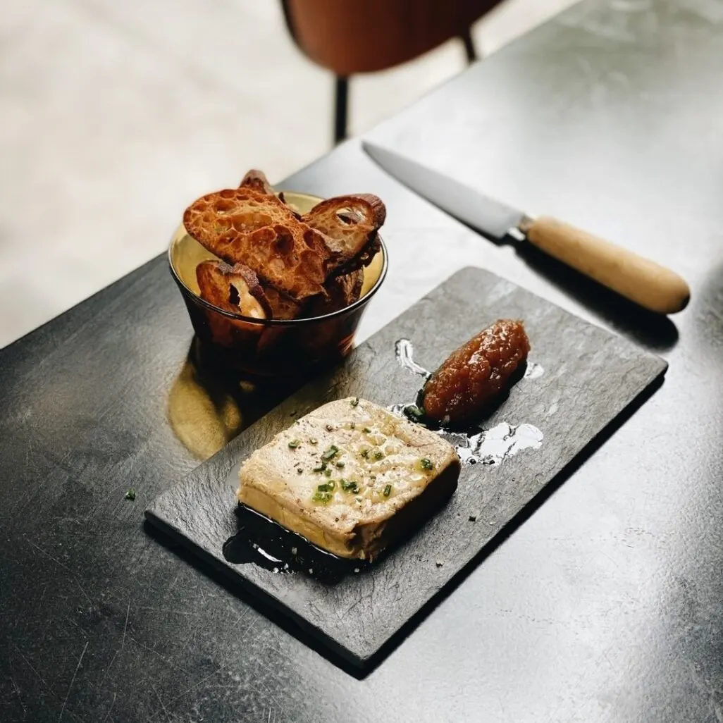 Tabla de pizarra con pâté, hierbas y chutney; al lado, cuenco con pan tostado y cuchillo de mango de madera sobre mesa oscura.