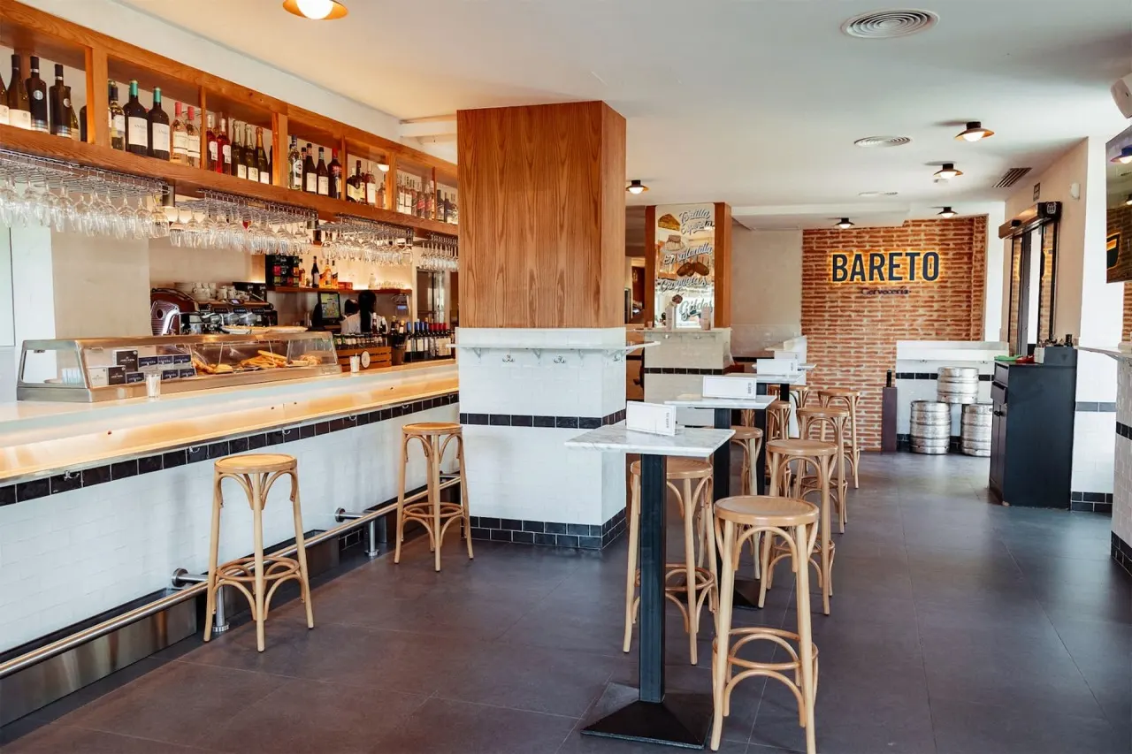 ChatGPT Plus Interior de Bareto Olavide con barra de madera, botellas expuestas y mesas altas, todo en un ambiente castizo y moderno con paredes de ladrillo visto.