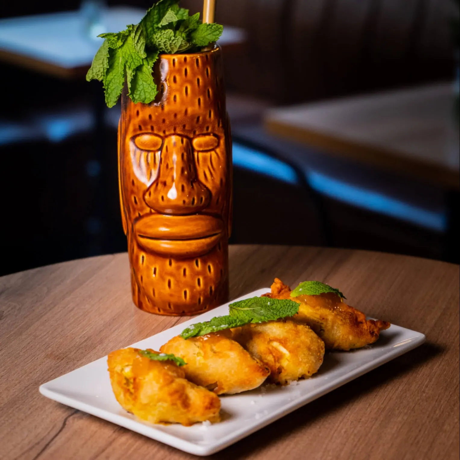 Plato con empanadas doradas decoradas con menta junto a cóctel en vaso tiki con hojas frescas, sobre mesa de madera en ambiente acogedor.
