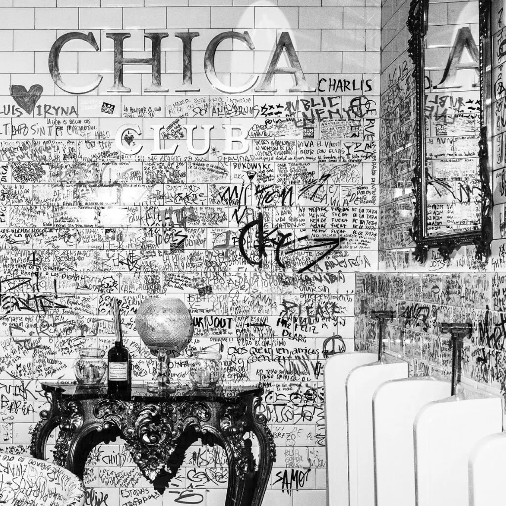 Sala con paredes de azulejos blancos llenas de grafitis, mesa tallada con globo y vino; texto “CHICA CLUB” destacado arriba.
