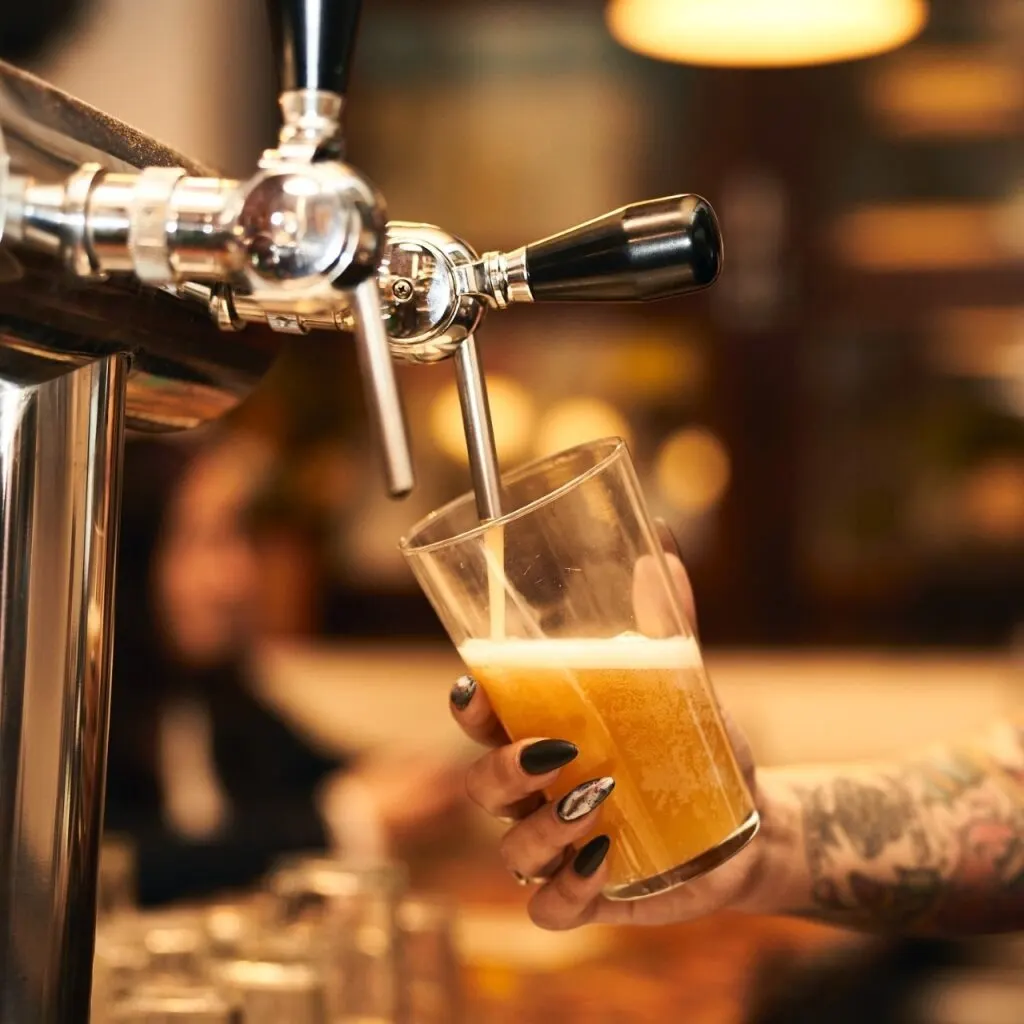 Mano con uñas negras y tatuajes sirve cerveza ámbar con espuma desde grifo metálico en bar con luces cálidas y fondo desenfocado.