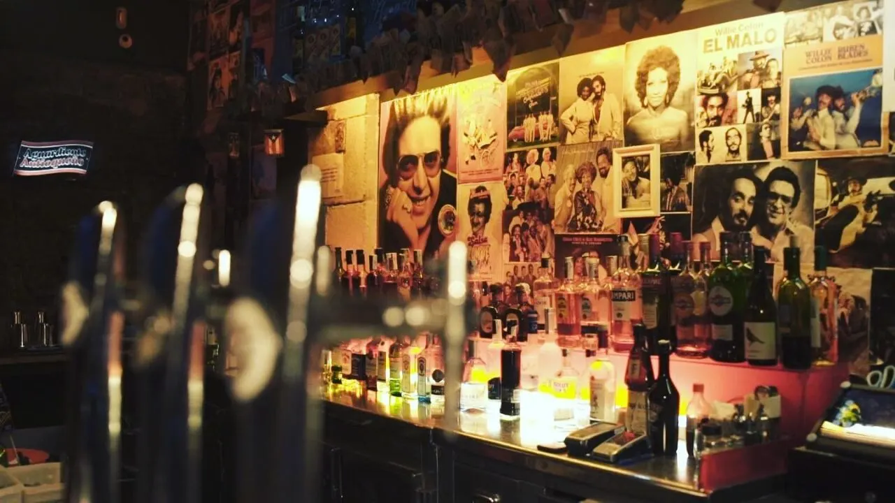 Interior de bar con botellas de licor en la barra, pared decorada con pósters de músicos y grifos de cerveza. Iluminación tenue y ambiente acogedor.