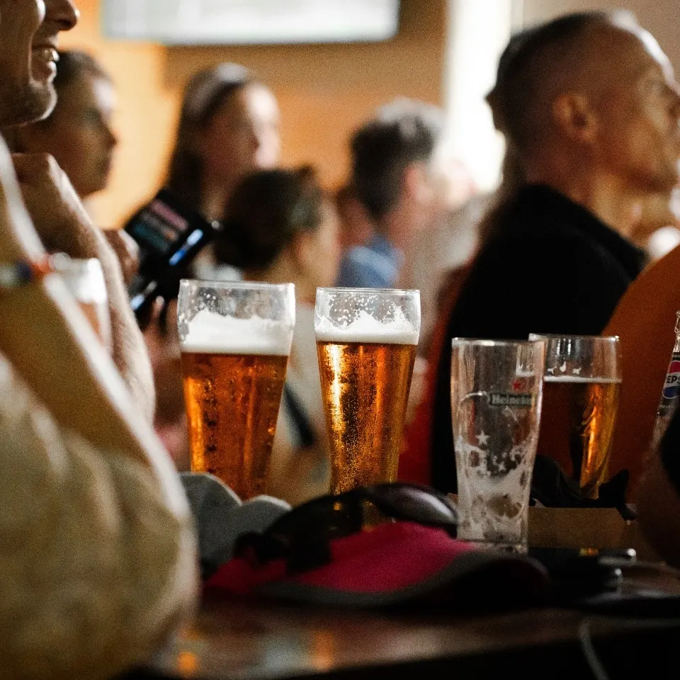 Copas de cerveza sobre la barra, con gente al fondo viendo una pantalla. Ambiente animado y distendido.