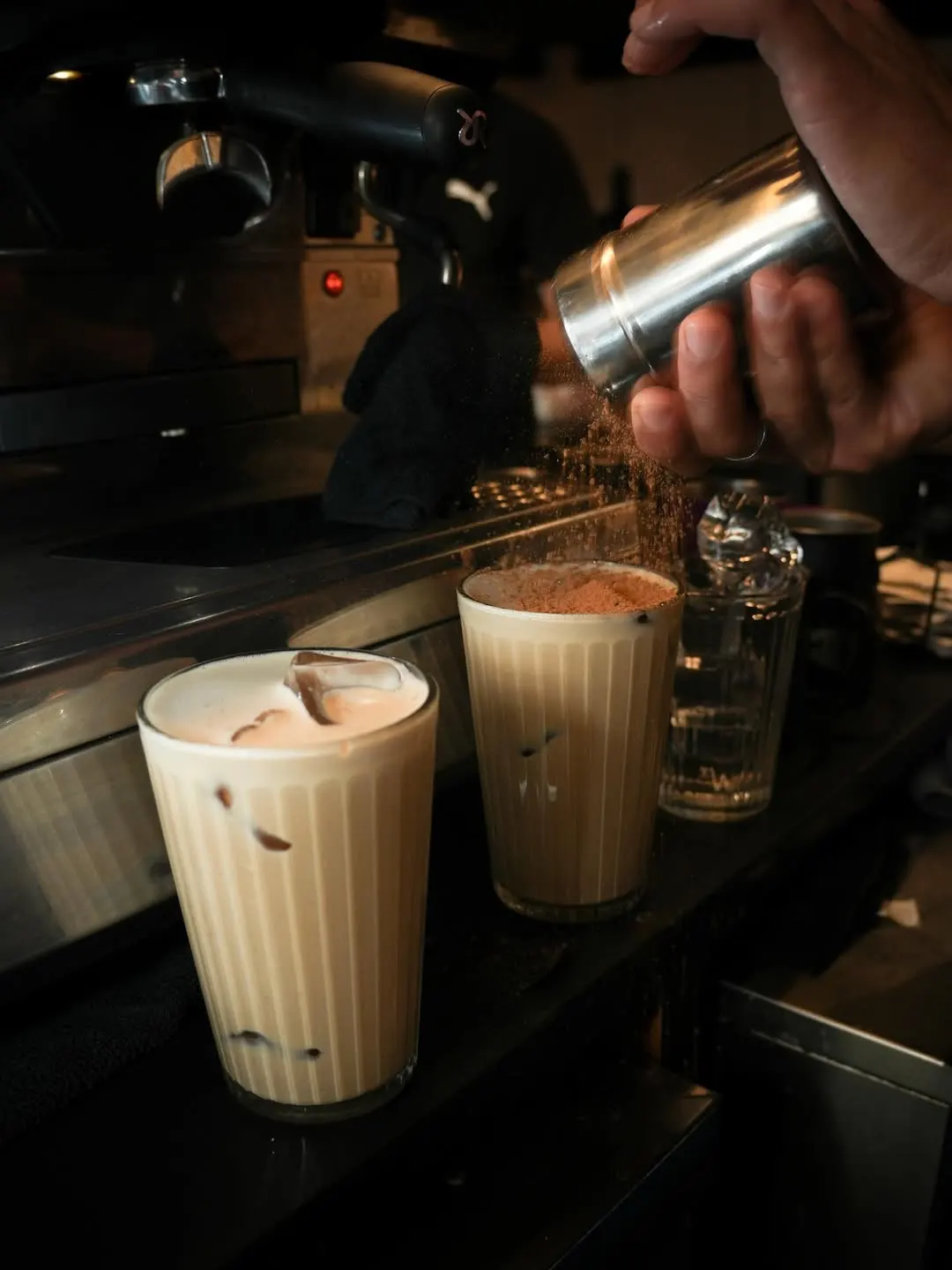 Dos vasos de café helado en God Café con hielo y espuma mientras se espolvorea cacao sobre la superficie junto a la máquina de café