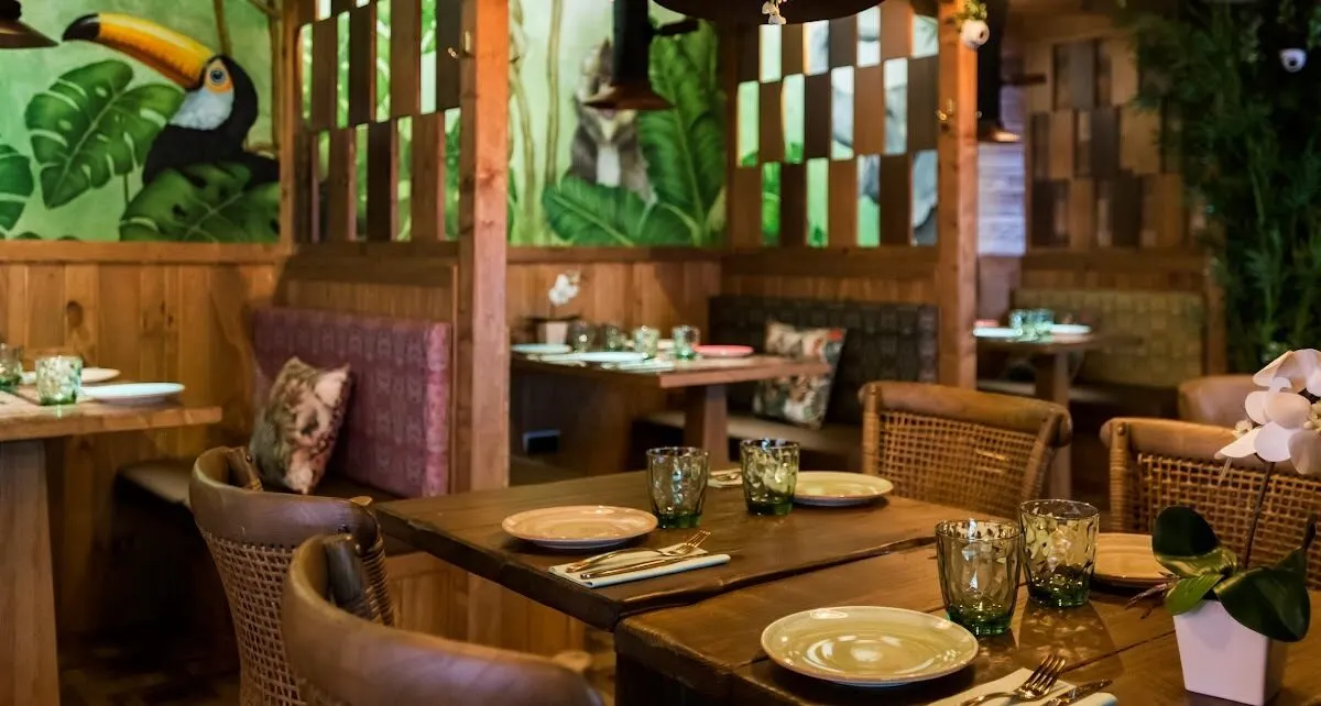 Interior de restaurante con muebles de madera, sillas de mimbre y mural tropical con hojas y animales. Mesas puestas y ambiente exótico y acogedor.