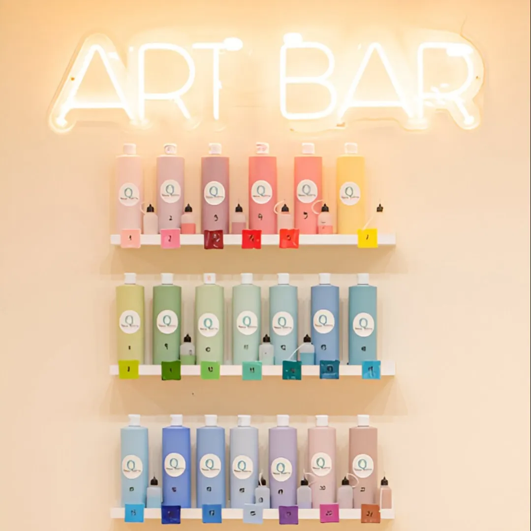 Pared de tonos suaves en Mama Pottery con estanterías llenas de botes de pintura ordenados por color bajo un luminoso letrero de neón que dice "ART BAR".