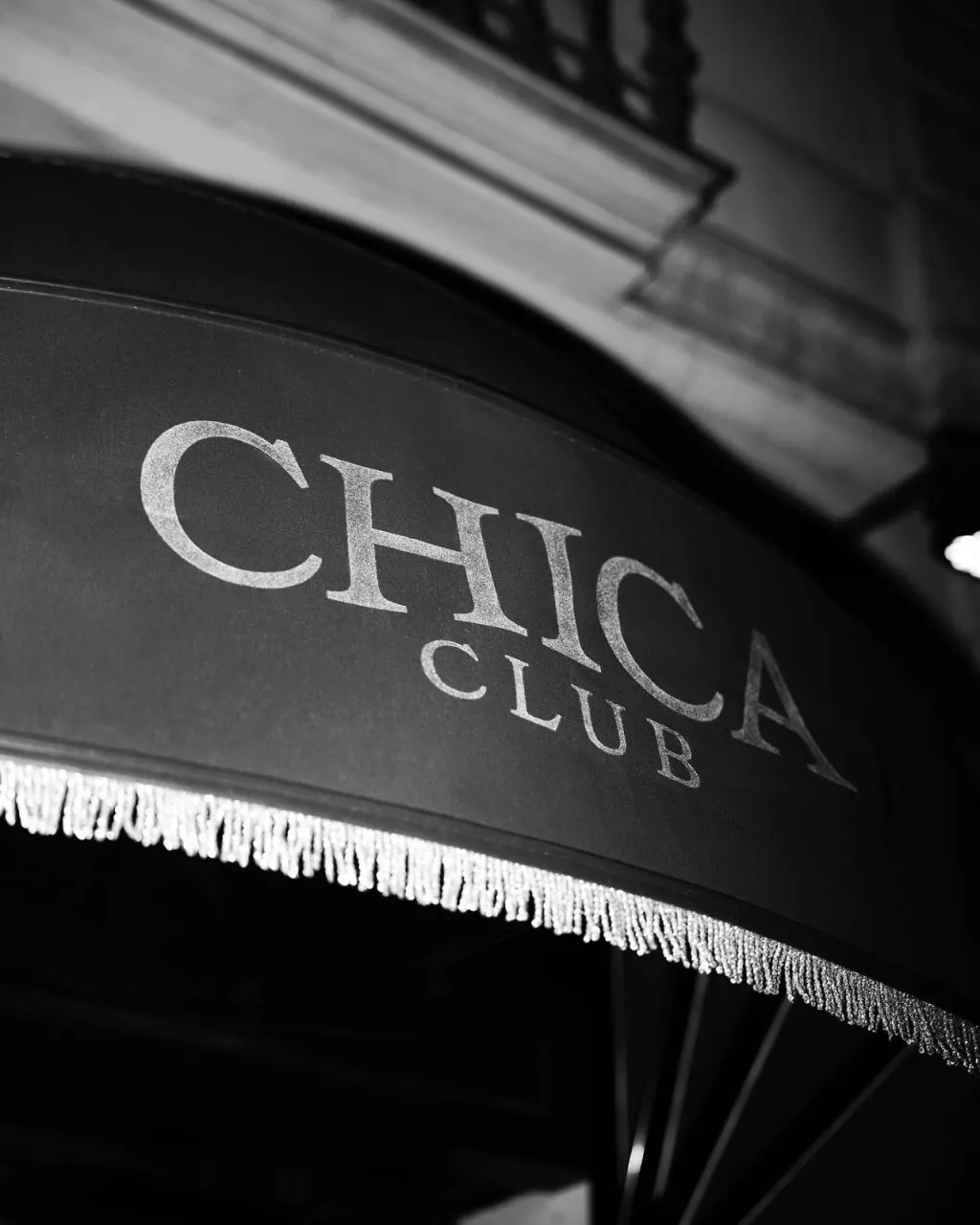 Fotografía en blanco y negro de un toldo con flecos y el texto “CHICA CLUB”. Estilo sencillo y elegante.
