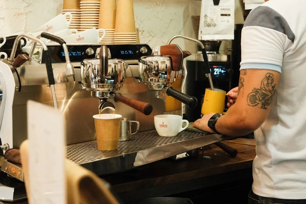 Barista preparando café con máquina profesional, vasos de cartón, tazas de cerámica y leche vaporizada en una cafetería de estilo moderno.