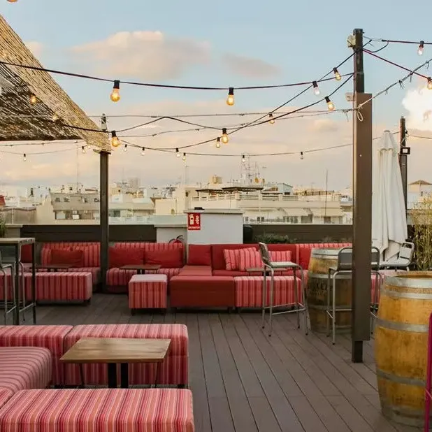 Terraza con sofás a rayas rojas y blancas, barriles como mesas, luces colgantes, sombrillas y vista urbana bajo cielo parcialmente nublado.
