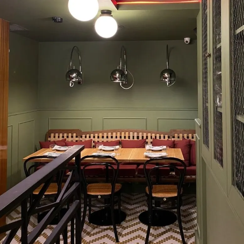 Comedor con mesa de madera, sillas negras trenzadas, banco con cojines rojos, suelo en zigzag y lámparas esféricas colgantes.