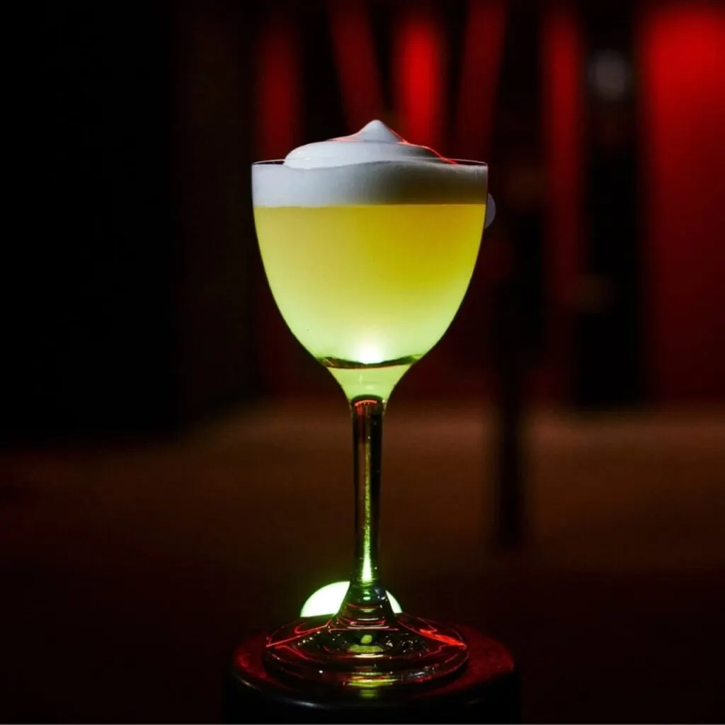Cóctel amarillo con espuma blanca en copa elegante; fondo oscuro con iluminación roja que resalta el contraste y la presentación del trago.