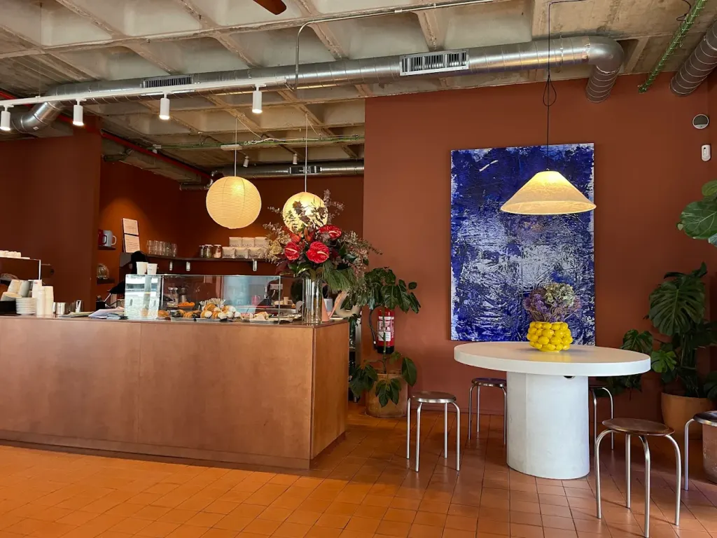 En la imagen se ve una cafetería con barra de madera, lámparas colgantes redondas, flores decorativas y una mesa blanca con limones frente a una pared color terracota.