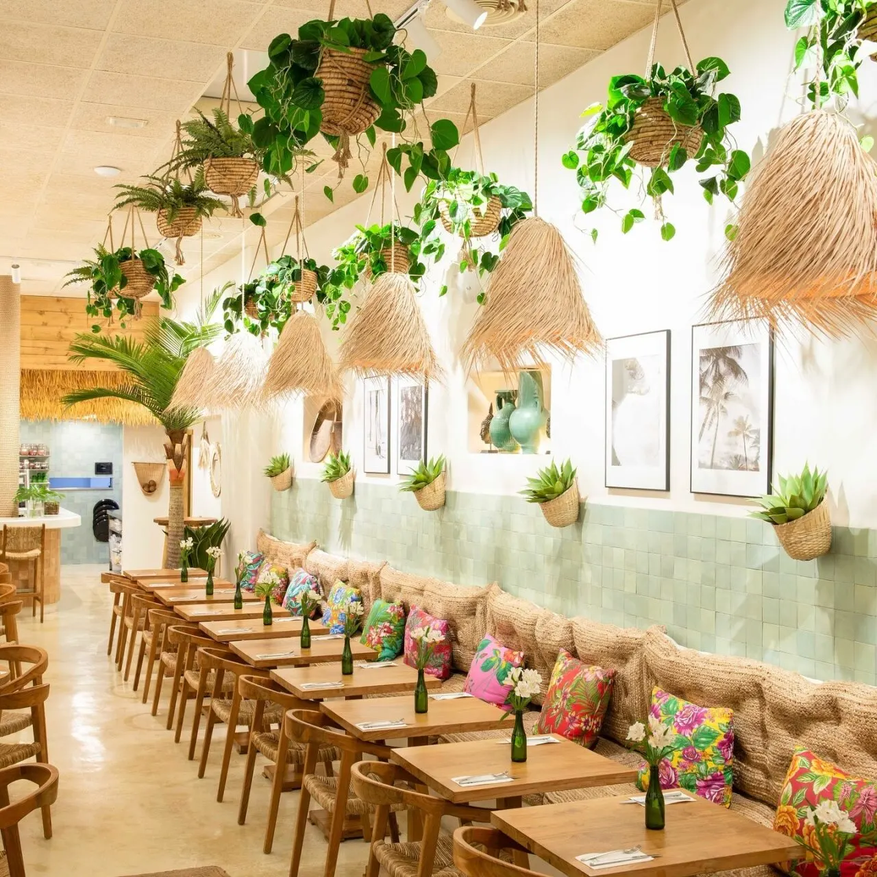 Zona de comedor interior con mesas de madera, cojines florales, plantas colgantes y decoración tropical. Ambiente luminoso y acogedor.