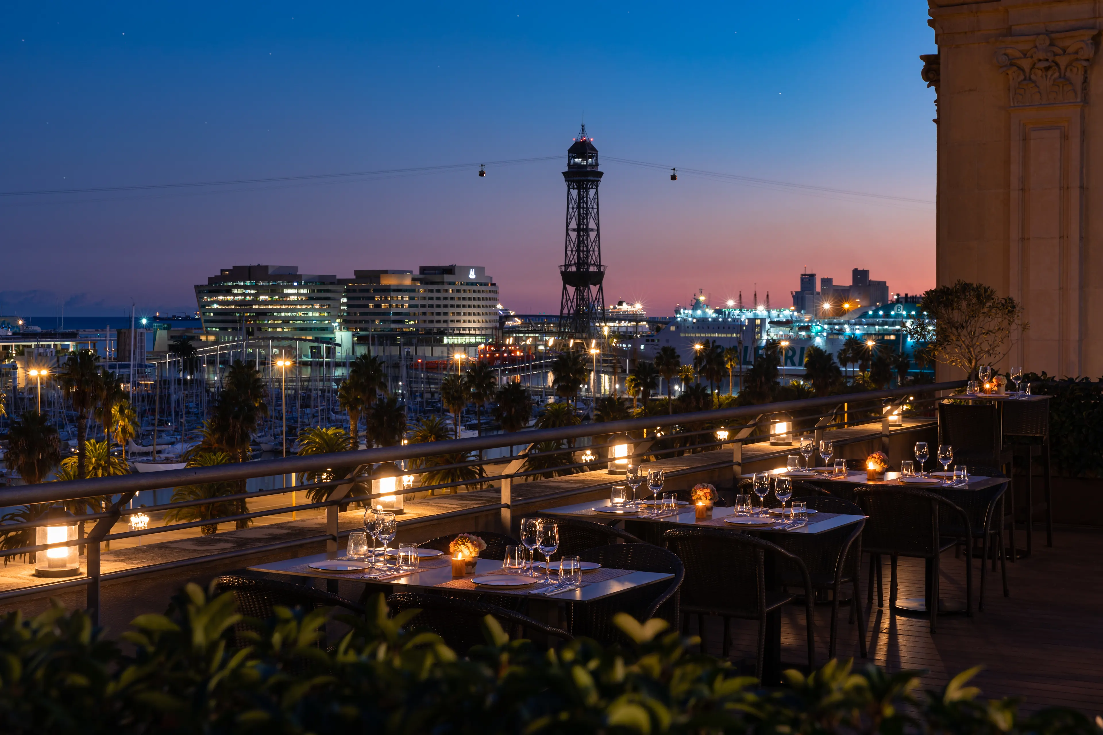 La foto muestra una terraza de comedor de La Terraza de la Duqesa, preparada para una comida con vista a un paisaje urbano durante el crepúsculo. Las mesas están adornadas con velas encendidas, copas de vino y servilletas