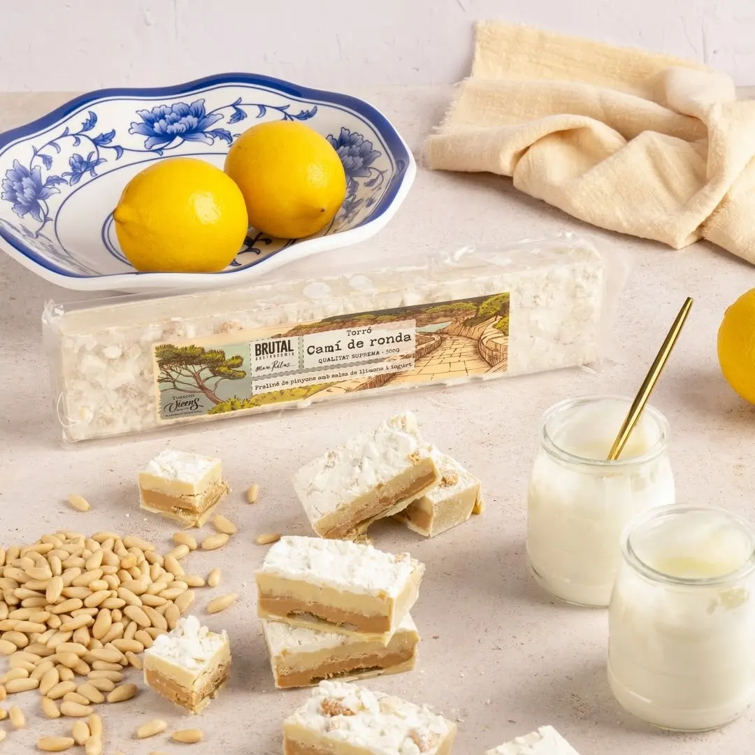 Trozos de turrón "Camí de Ronda" con piñones, limón y azúcar glas, presentados junto a piñones crudos, limones frescos, yogur y una fuente decorativa sobre fondo claro.