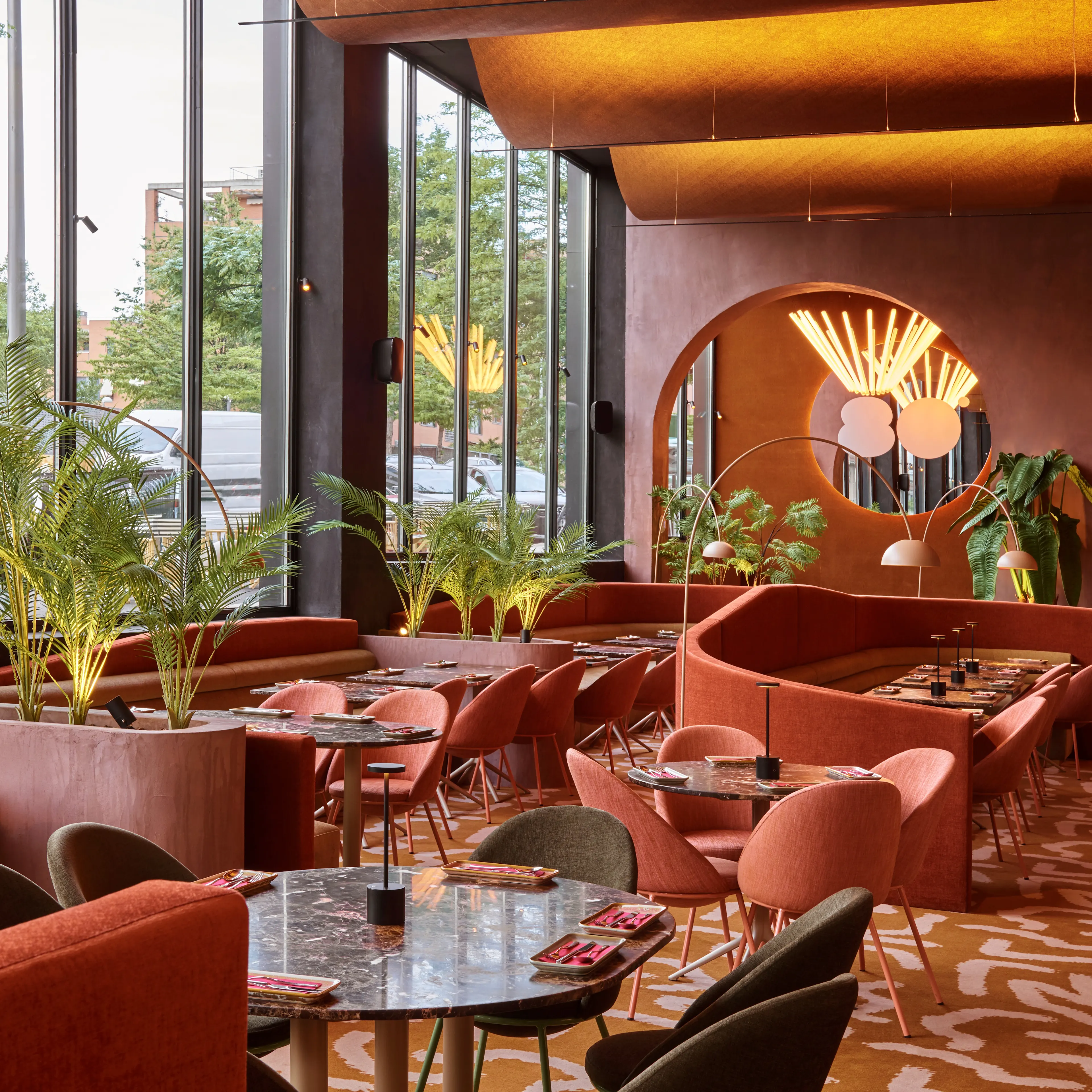 Interior de Ochenta Grados Montecarmelo con grandes ventanales, plantas, tonos cálidos y un diseño moderno con mesas redondas y sofás envolventes.