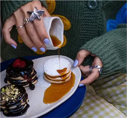 Persona con suéter verde vierte sirope sobre pancakes; hay otras dos torres con chocolate, frambuesas y pistachos. Uñas lilas y anillos visibles.